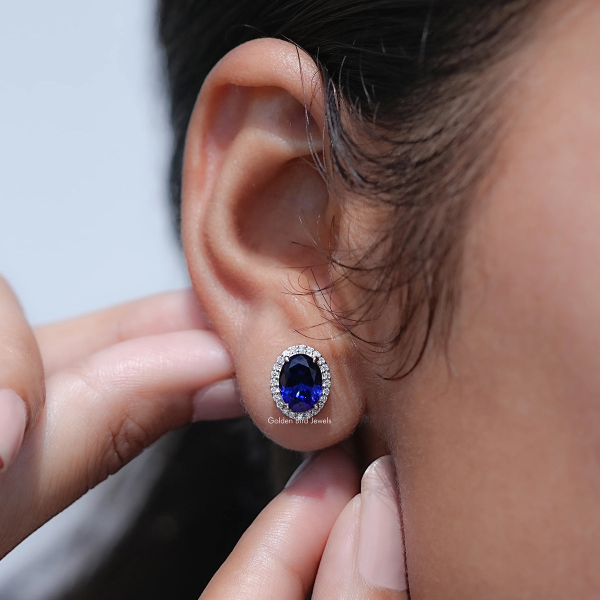 Oval blue gemstone halo stud earring, close-up on ear with round halo setting