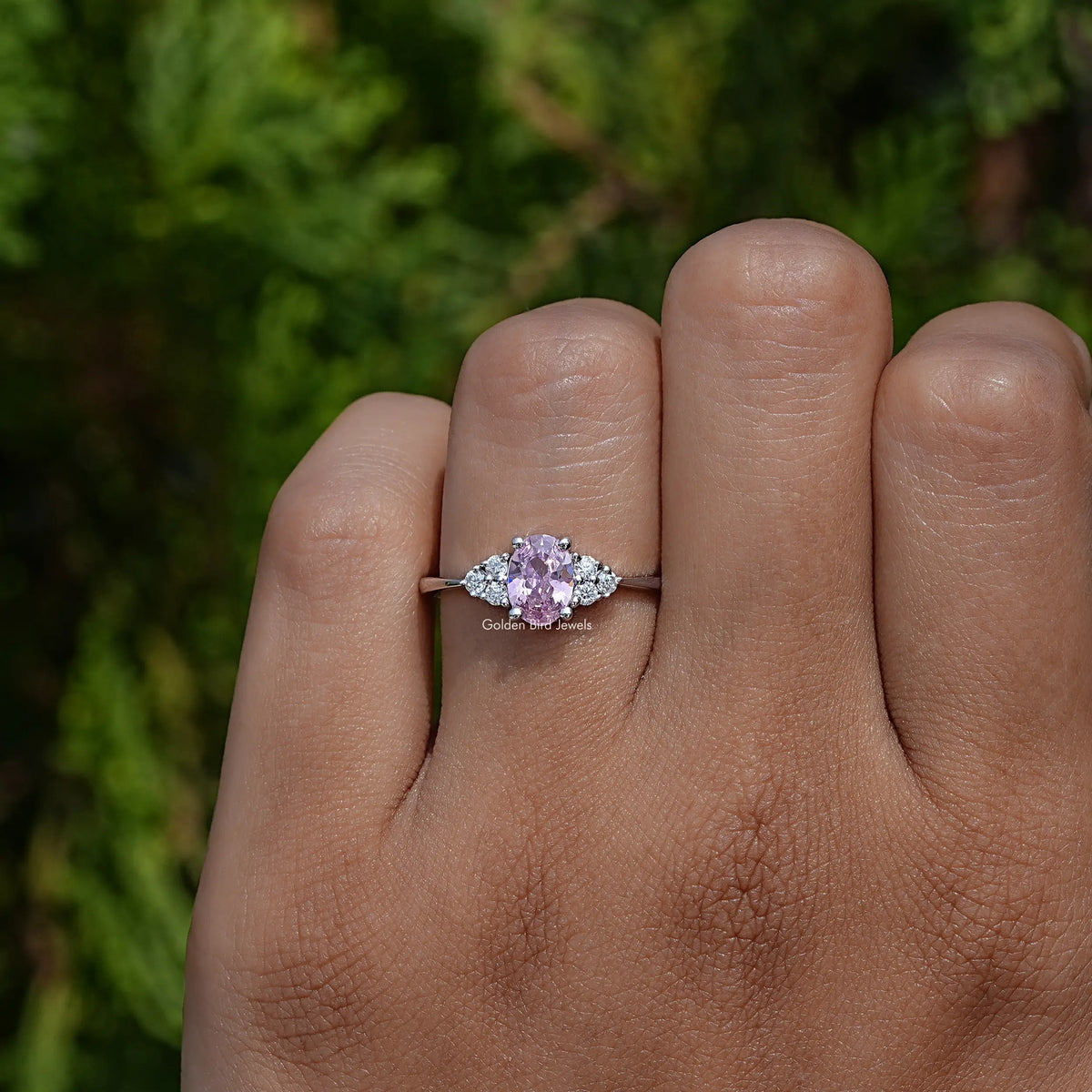 Oval pink gemstone engagement ring on palm with round side stones and slender band