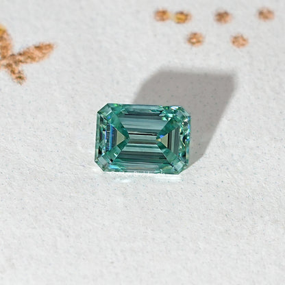 Emerald-cut teal-green moissanite loose stone centered on light surface