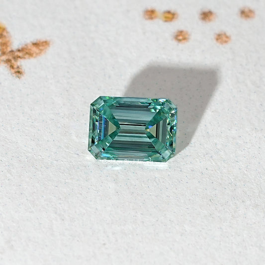 Emerald-cut teal-green moissanite loose stone centered on light surface