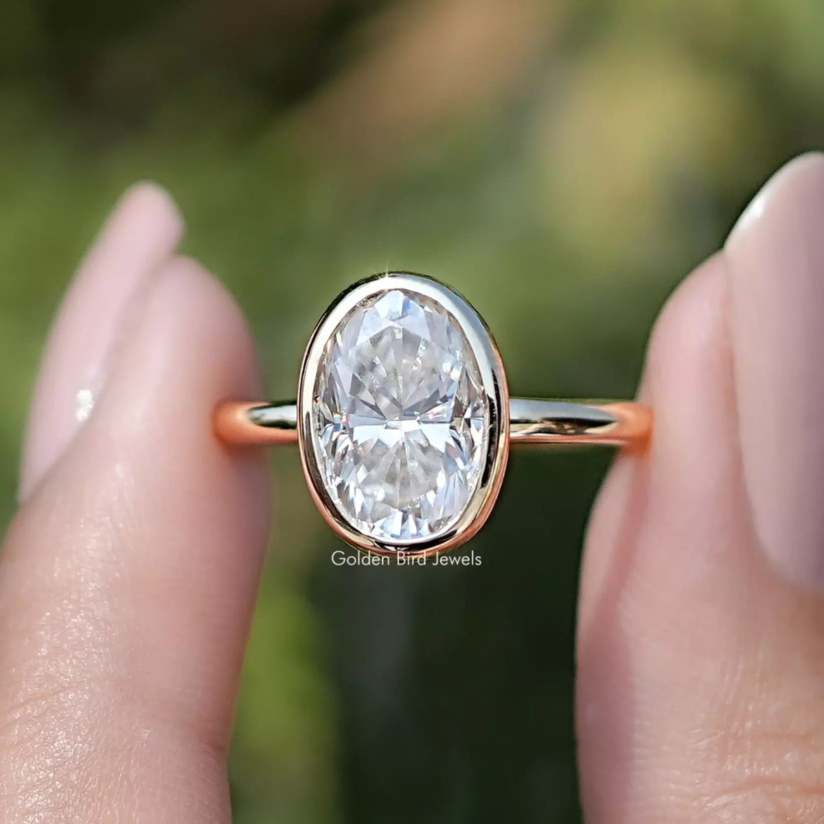Front view of Bezel Set Oval Moissanite Engagement Ring in two fingers 
