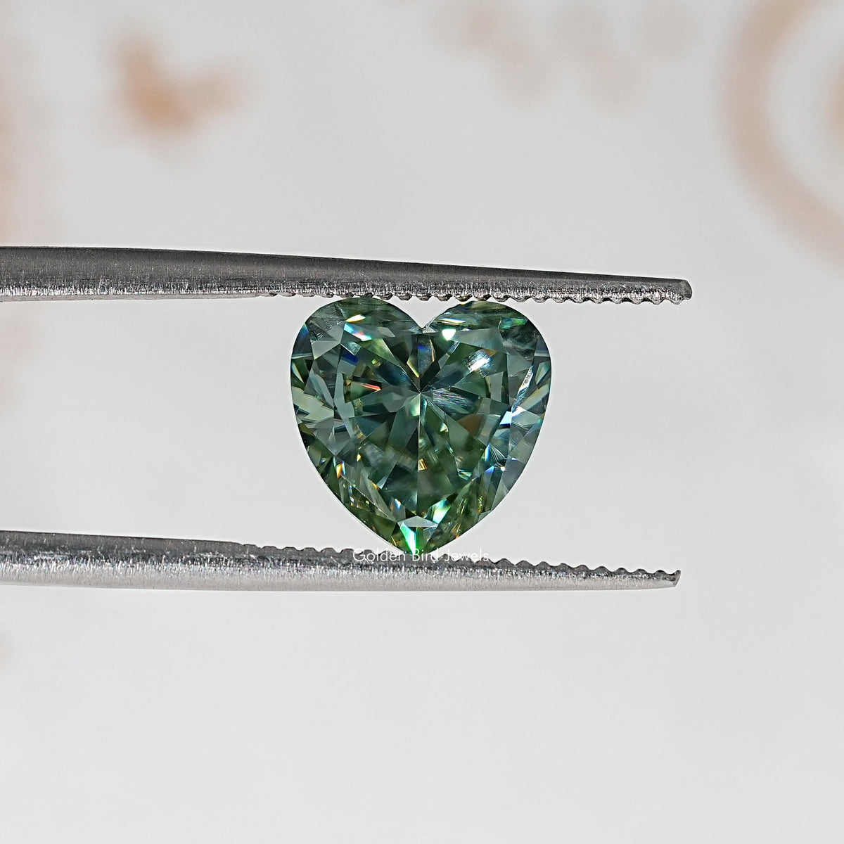 Heart-cut moissanite gemstone in tweezers, green-blue faceted surface.