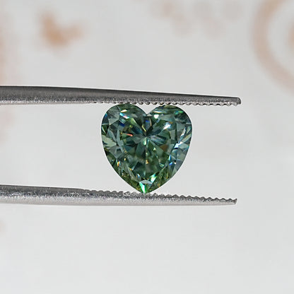 Heart-cut moissanite gemstone in tweezers, green-blue faceted surface.
