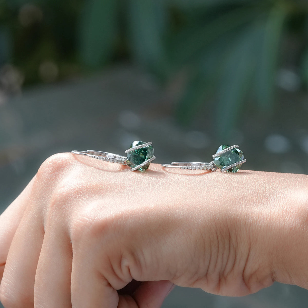 Two sterling silver dangle earrings with emerald-green cushion-cut moissanite on hand