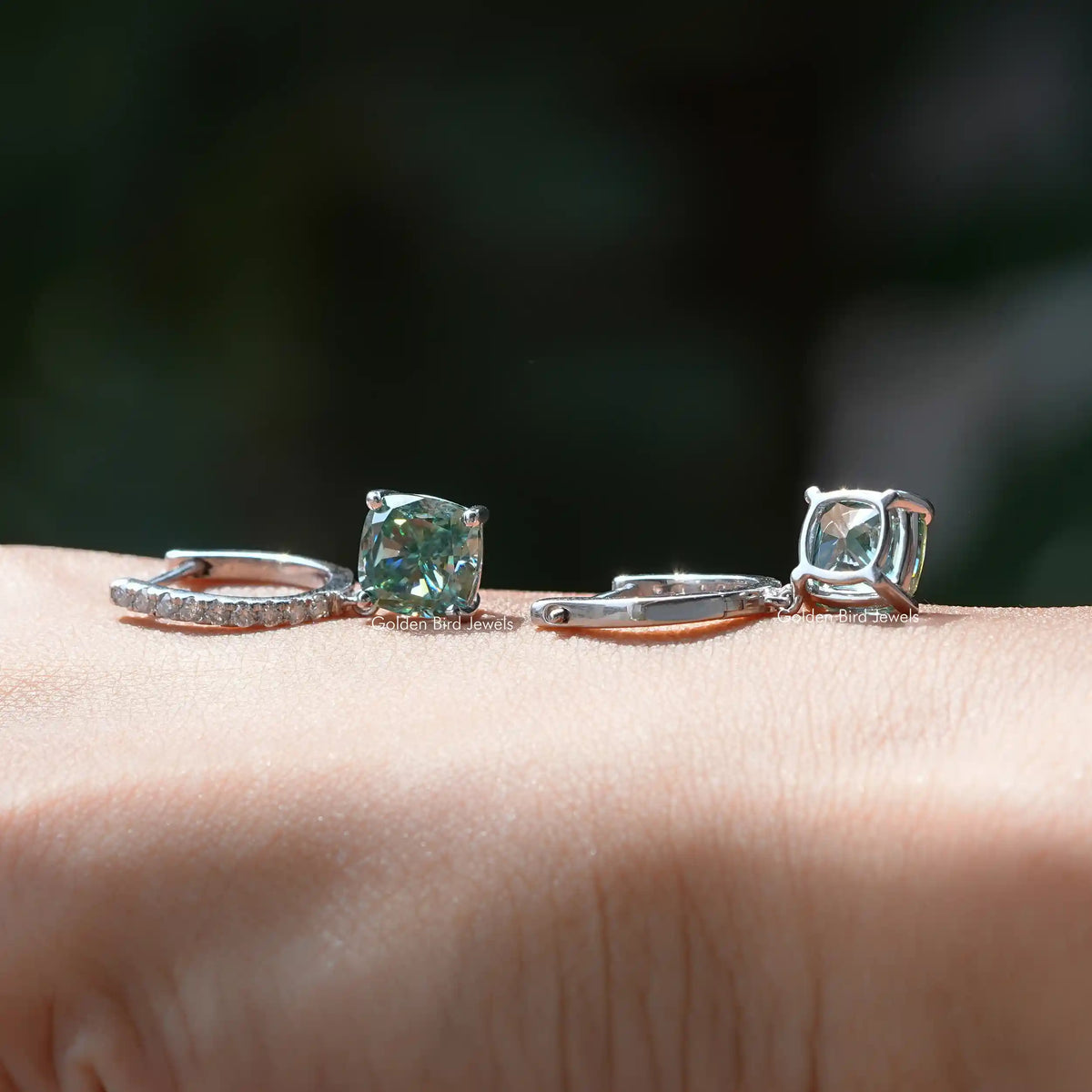 Cushion-cut blue green moissanite earrings in silver, shown on a wrist