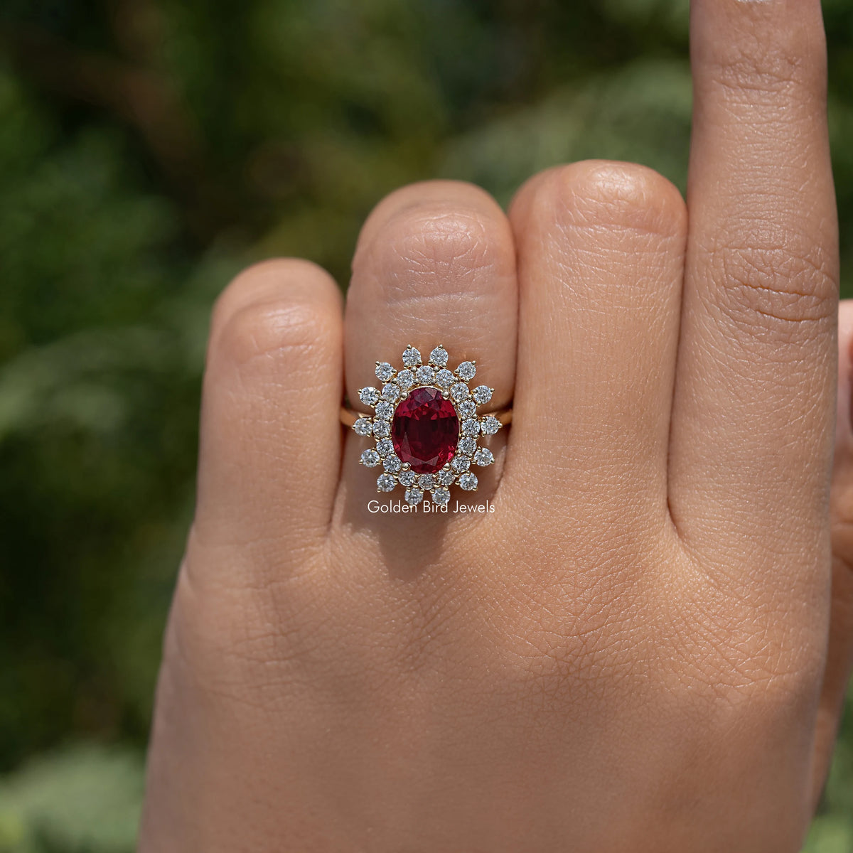 Oval red gemstone halo engagement ring with clustered round stones on finger