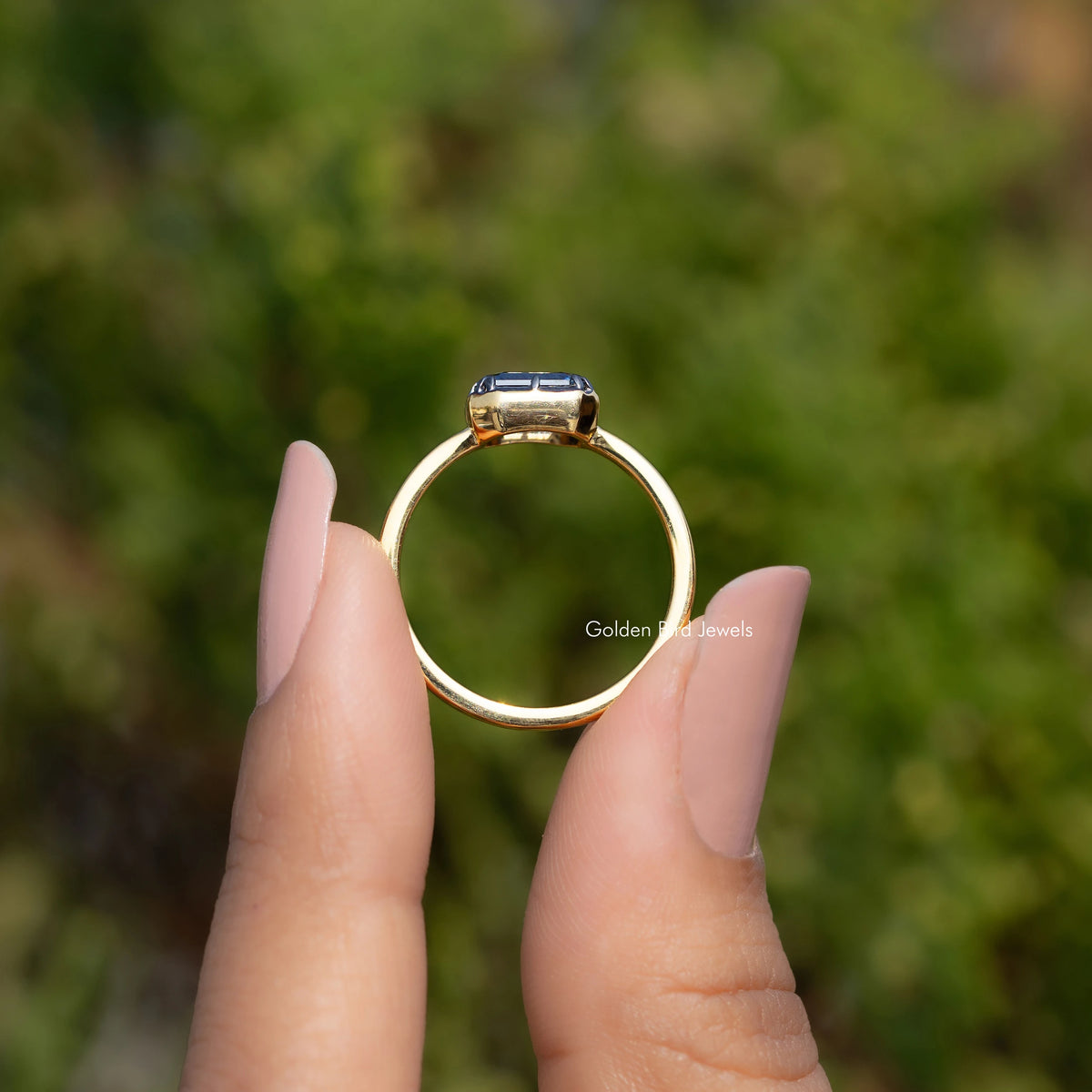 Gold solitaire ring with emerald-cut gemstone, side view held by fingers