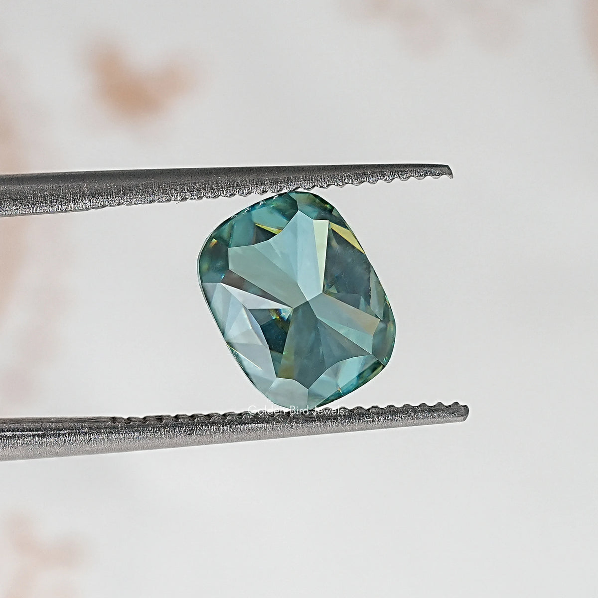 Elongated cushion teal-green moissanite stone between metal tweezers.