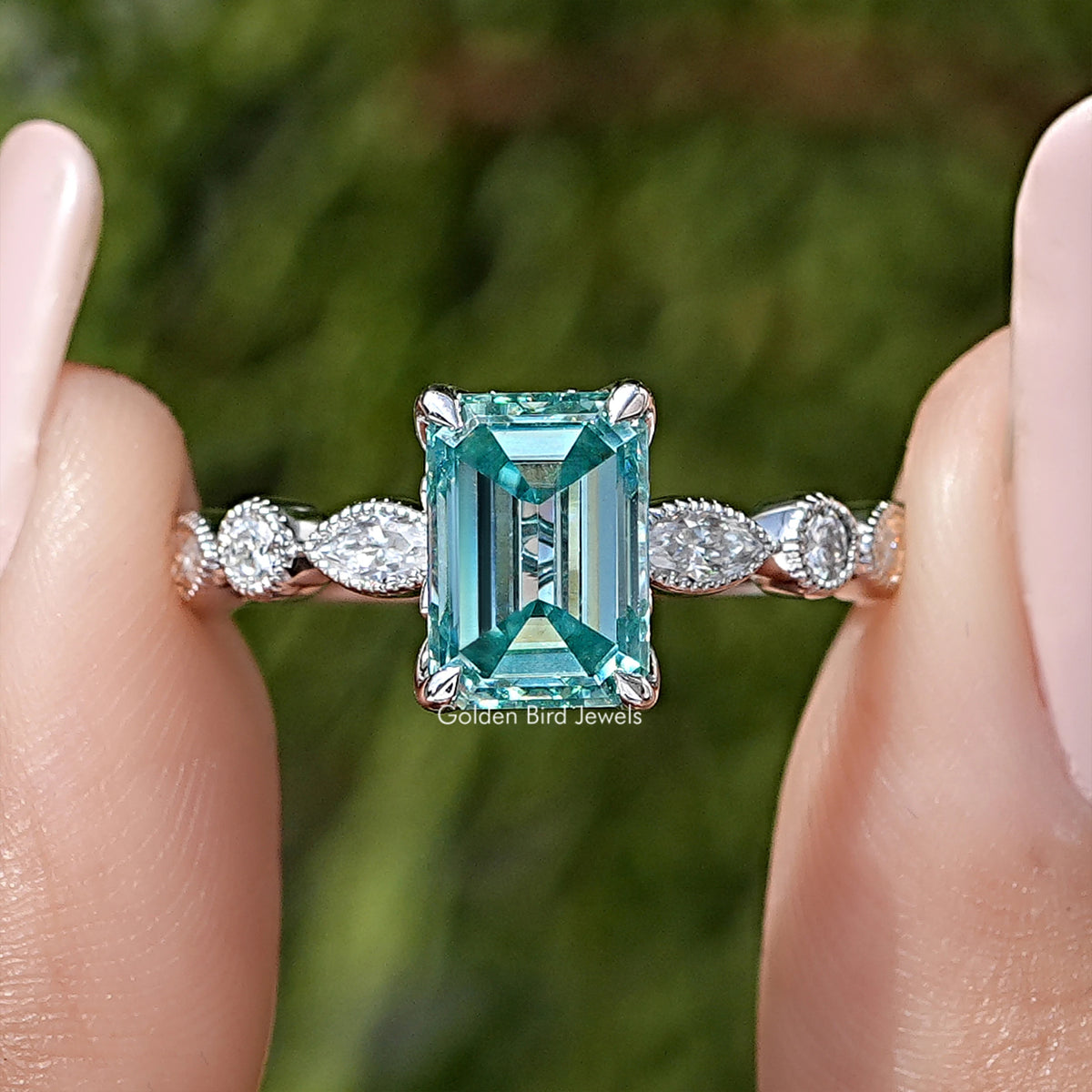 Emerald-cut moissanite engagement ring in silver, teal center with round side stones.