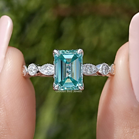 Emerald-cut moissanite engagement ring in silver, teal center with round side stones.