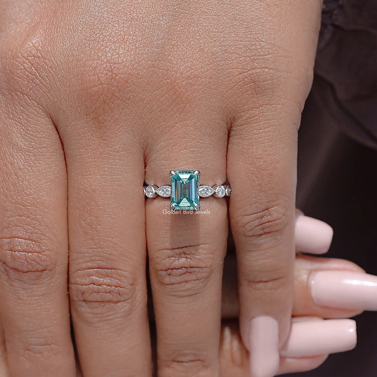 Emerald-cut moissanite ring with teal center and small round side stones on hand.
