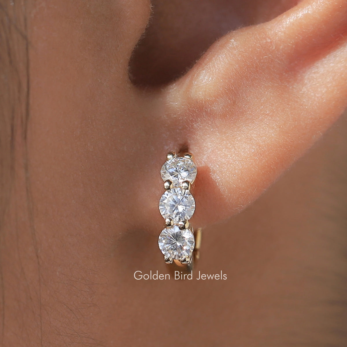 Gold-tone huggie hoop earrings with three round-cut stones, close-up on ear