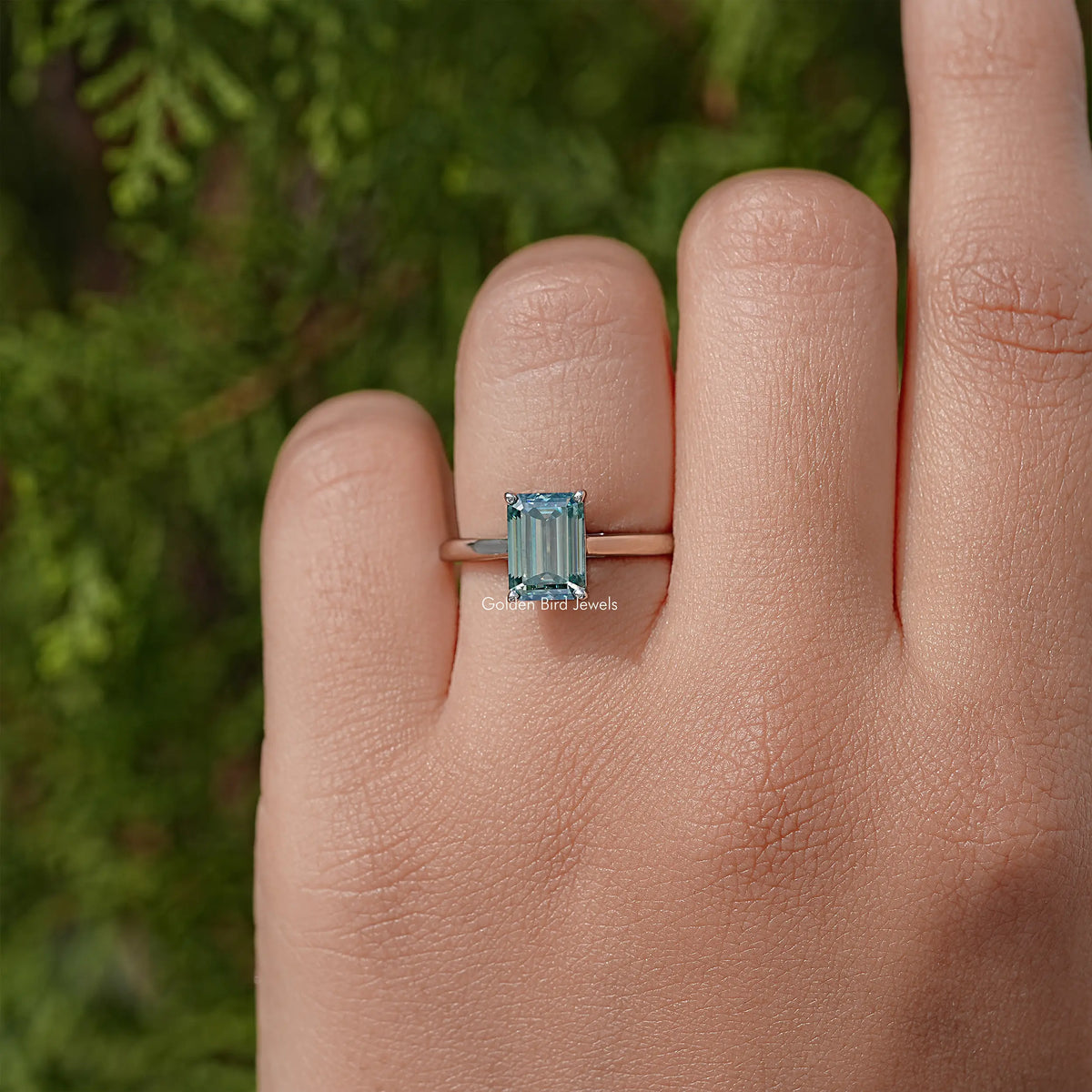 Emerald-cut moissanite solitaire ring on finger, square teal stone in silver setting