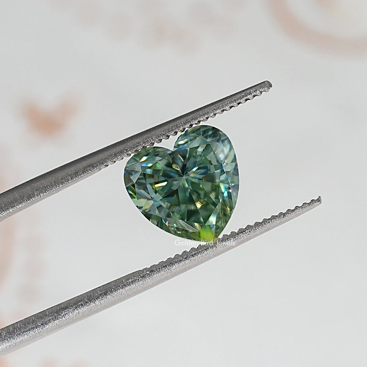 Heart-cut moissanite held between metal tweezers; green-blue facets visible.