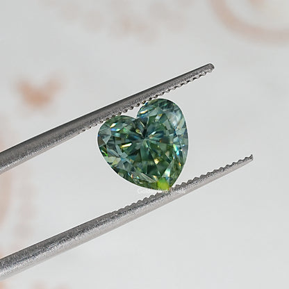 Heart-cut moissanite held between metal tweezers; green-blue facets visible.