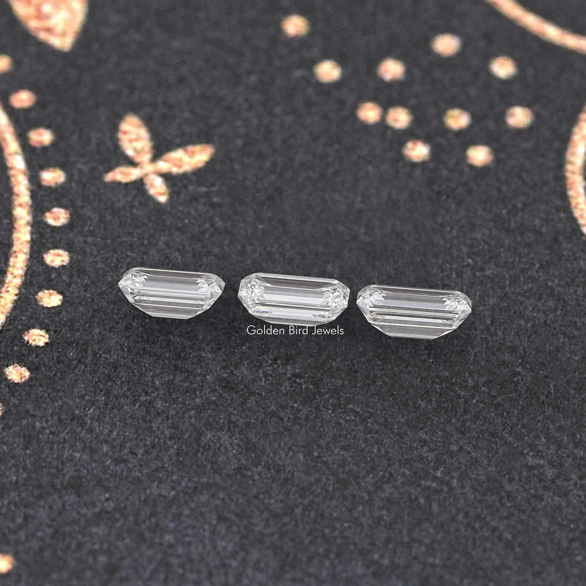 Three emerald-cut lab diamond stones, clear, lined up on dark fabric.