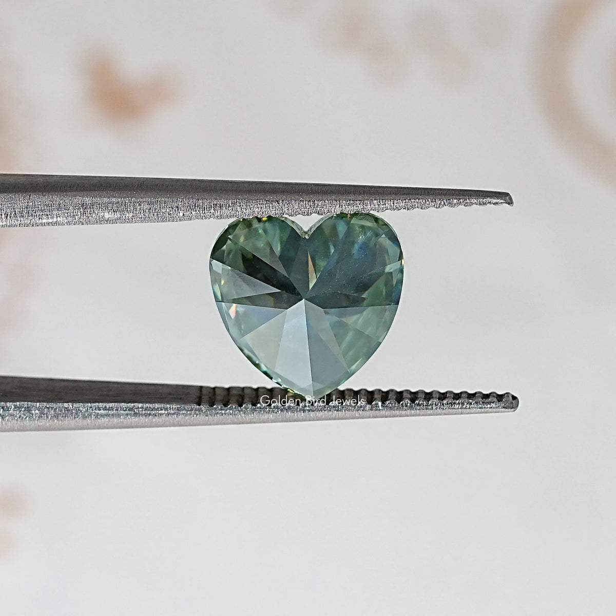 Heart-cut moissanite gemstone between metal tweezers; green-blue brilliance.