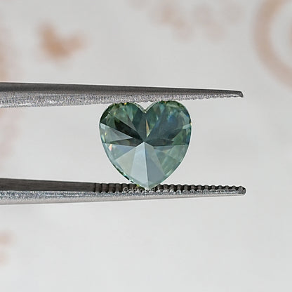 Heart-cut moissanite gemstone between metal tweezers; green-blue brilliance.
