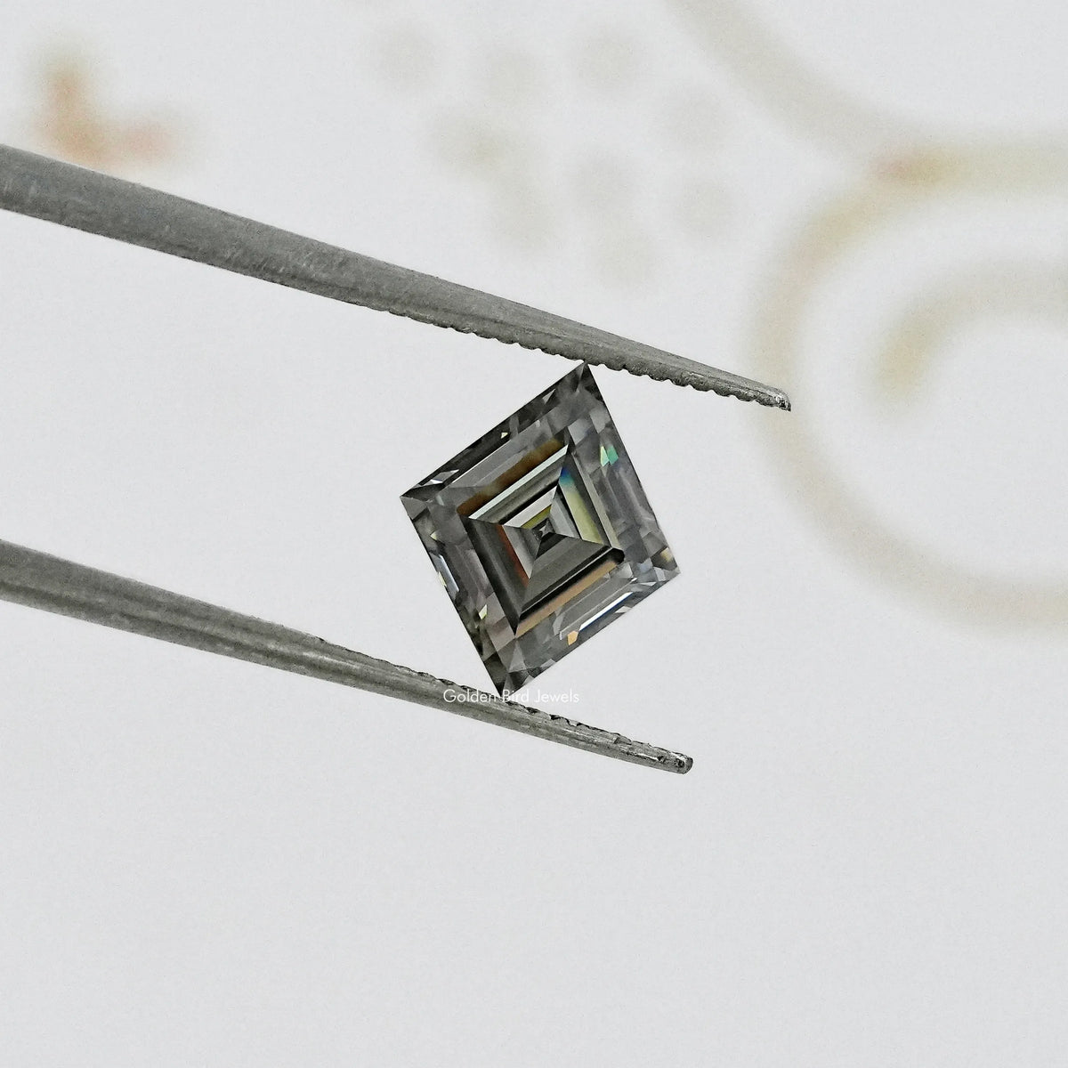 Gray lozenge-cut moissanite with step facets, held between metal tweezers.