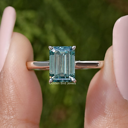 Emerald-cut moissanite solitaire ring close-up, teal gemstone with metal band and prongs