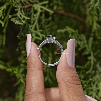 Sterling silver split-shank ring with heart-cut teal moissanite, held between fingers