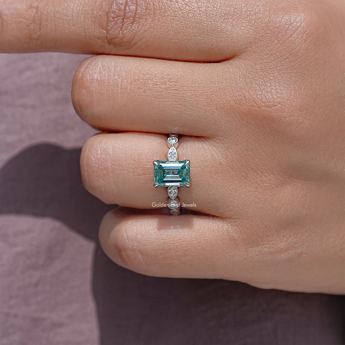 Silver emerald-cut moissanite ring on fingertip, teal center stone in prongs.