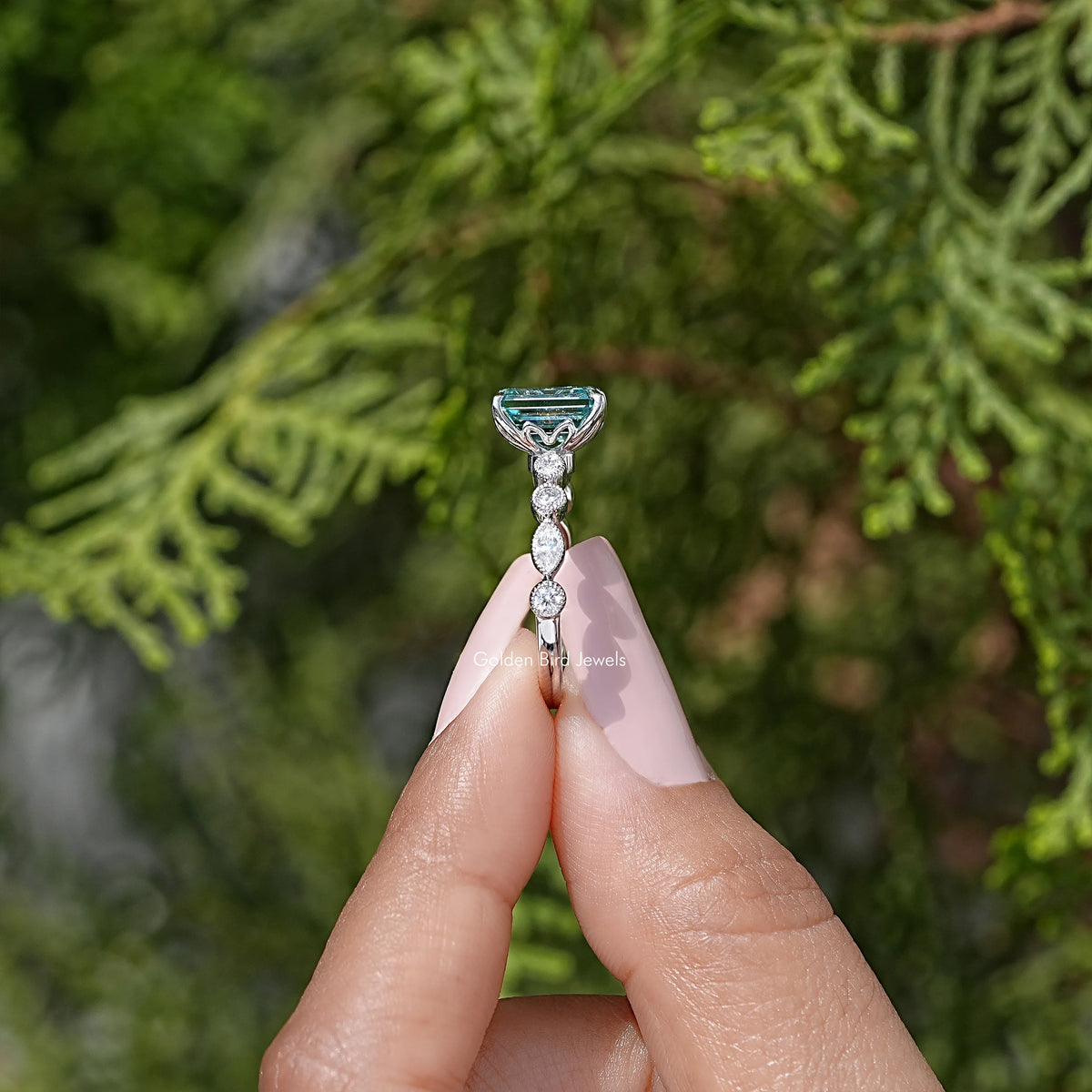 Emerald-cut moissanite ring top view: teal stone in prongs with small halo.