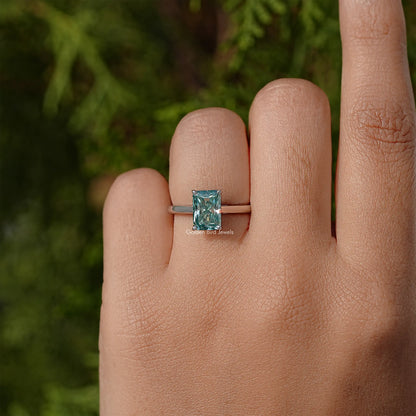 Sterling silver solitaire ring with emerald-cut blue-green moissanite on finger.