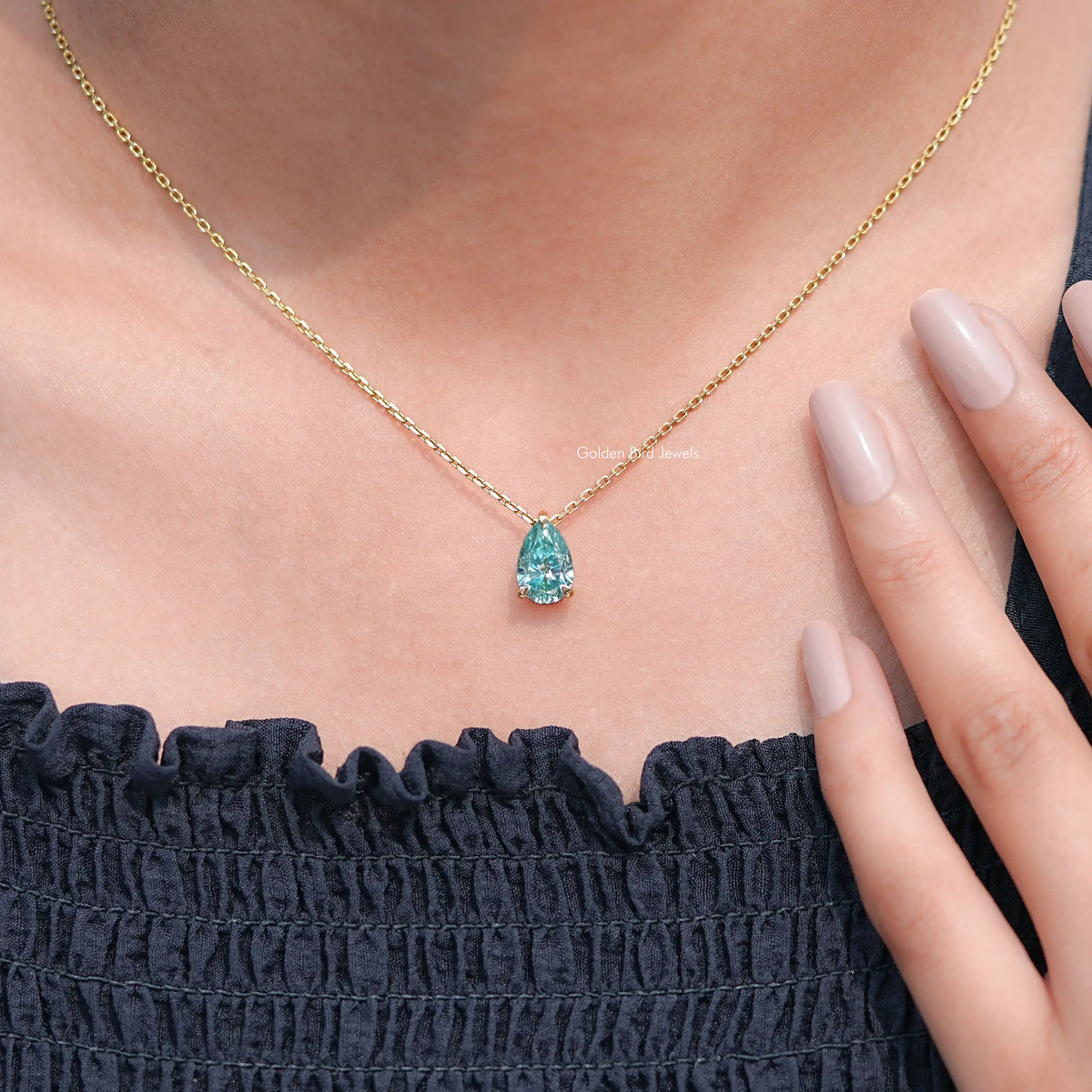 Gold chain pendant with pear-cut teal teardrop gemstone, worn with dark dress.