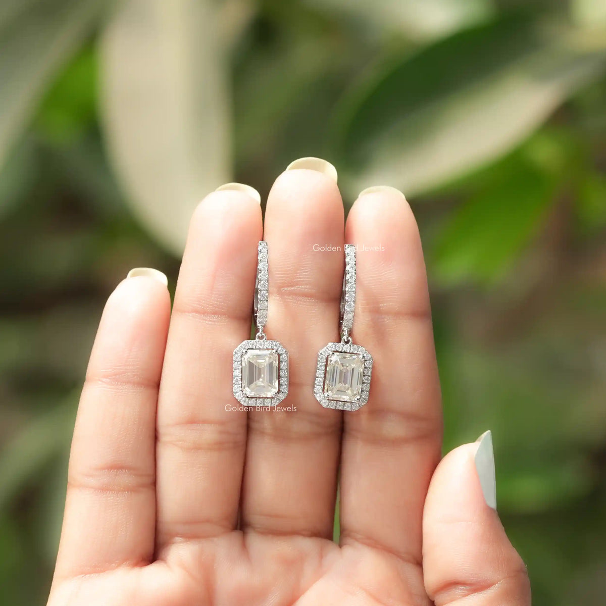 Pair of silver emerald-cut halo drop earrings shown in open hands