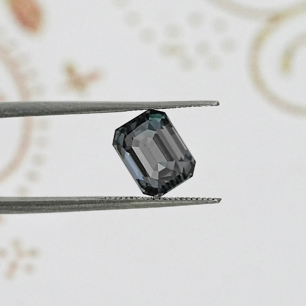 Gray emerald-cut gemstone close-up in tweezers, step facets visible