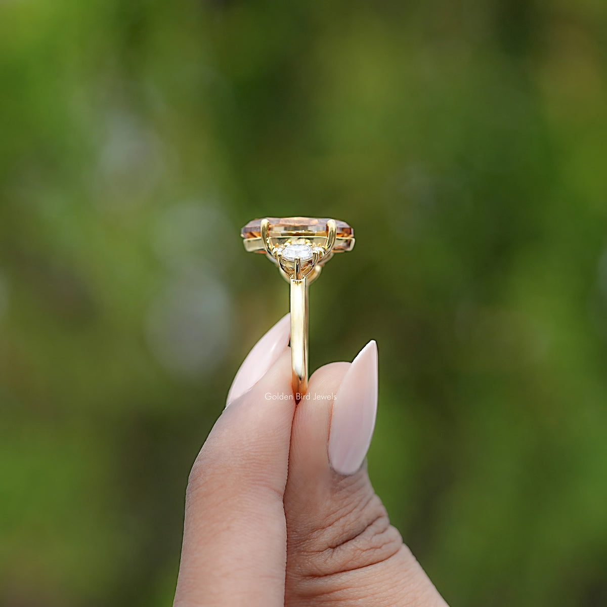 Moissanite engagement ring held up, oval amber center with round side stones, gold tone