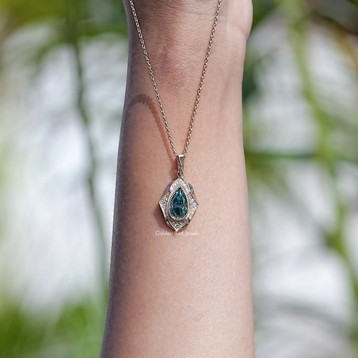 Pear-cut teal moissanite pendant necklace on wrist with gold-tone chain