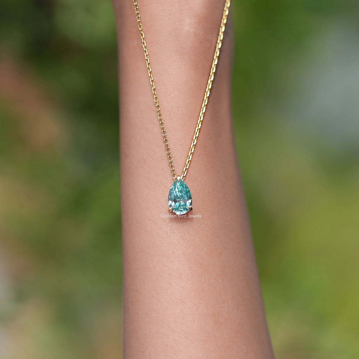 Gold chain pendant with pear-cut teal teardrop gemstone, worn on wrist.