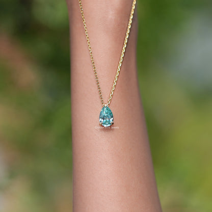 Gold chain pendant with pear-cut teal teardrop gemstone, worn on wrist.