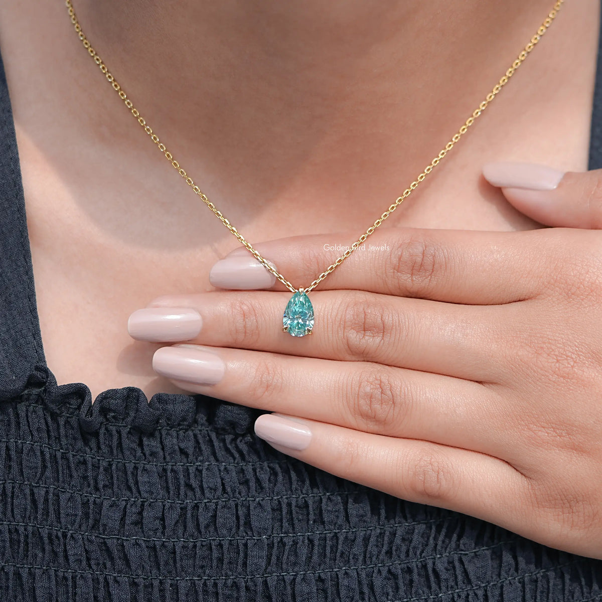 Gold chain pendant with pear-cut teal teardrop gemstone, held over dark dress.