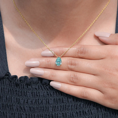 Gold chain pendant with pear-cut teal teardrop gemstone, held over dark dress.
