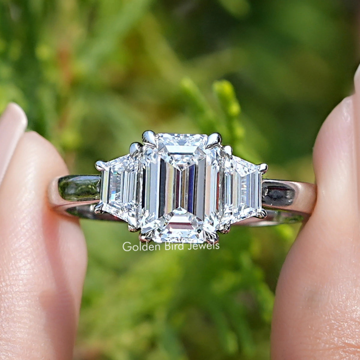 Three Stone Emerald Cut Diamond Engagement Ring Held Between Fingers