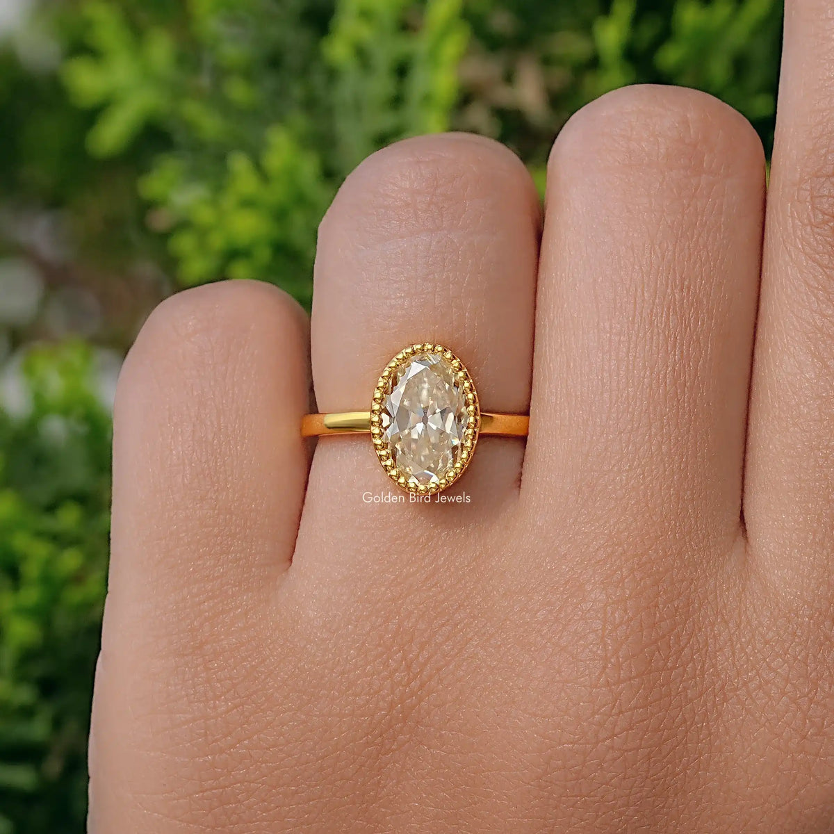 Gold-tone oval moissanite ring with milgrain bezel, worn on hand.