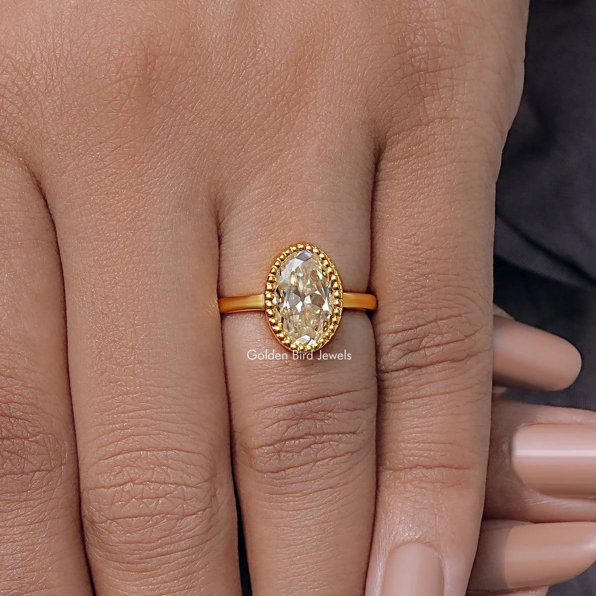 Oval moissanite ring in gold tone with milgrain bezel, worn on finger.