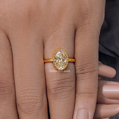 Oval moissanite ring in gold tone with milgrain bezel, worn on finger.