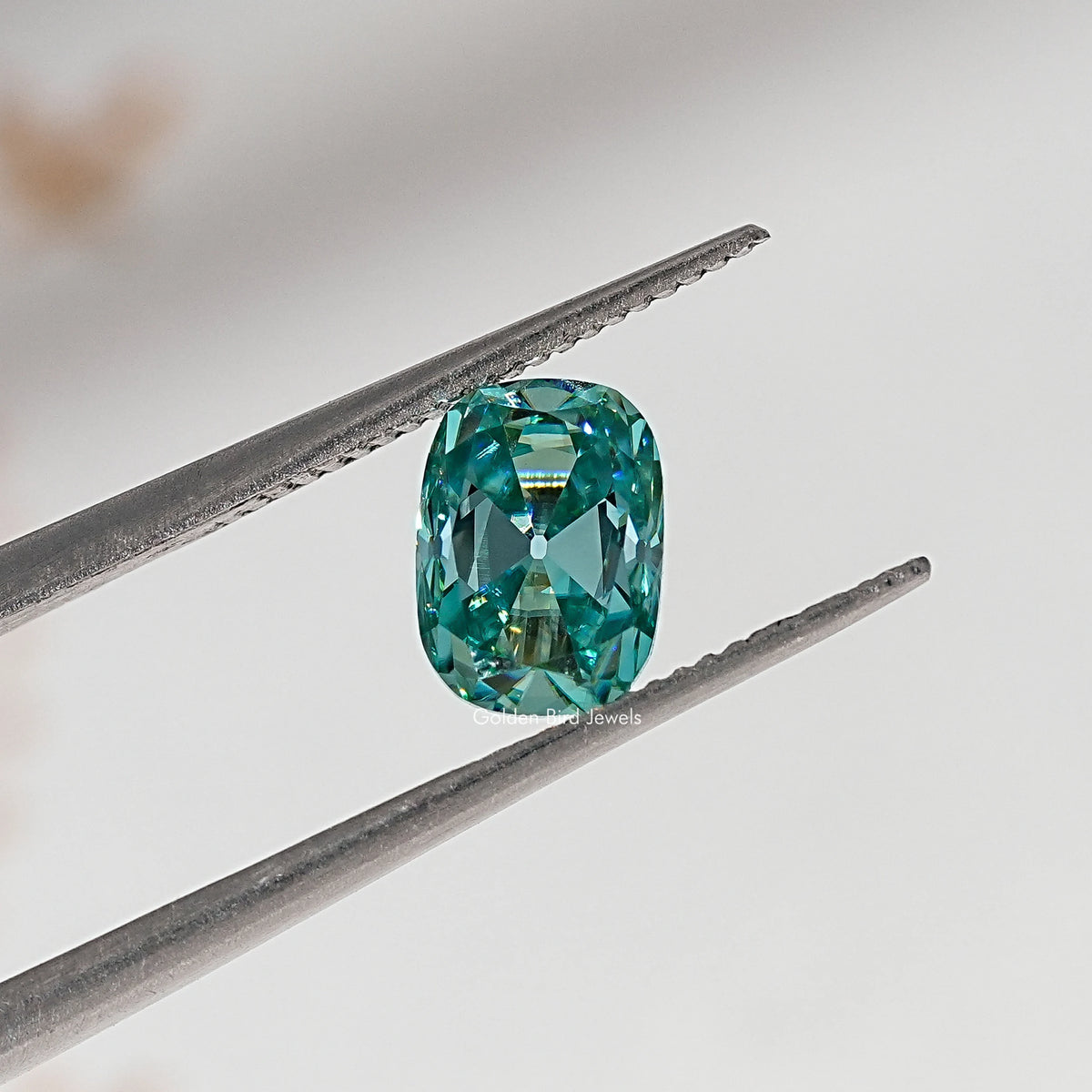 Blue-green cushion-cut moissanite gemstone held in tweezers, close-up facet view.