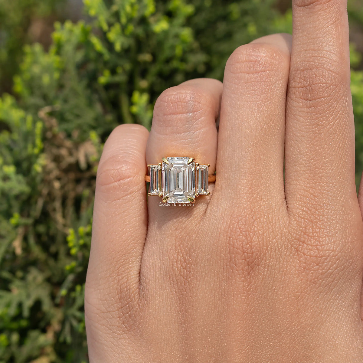 Three-stone emerald-cut moissanite engagement ring on hand, gold-tone band and square stones.