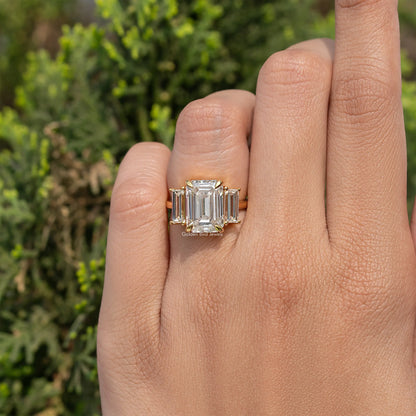 Three-stone emerald-cut moissanite engagement ring on hand, gold-tone band and square stones.
