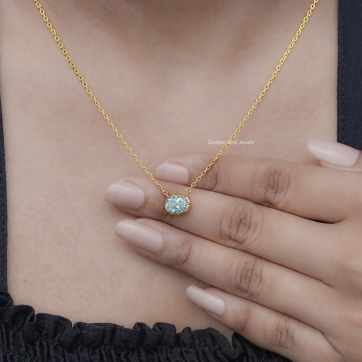 Oval moissanite solitaire pendant on gold-tone chain held in hand; blue gemstone