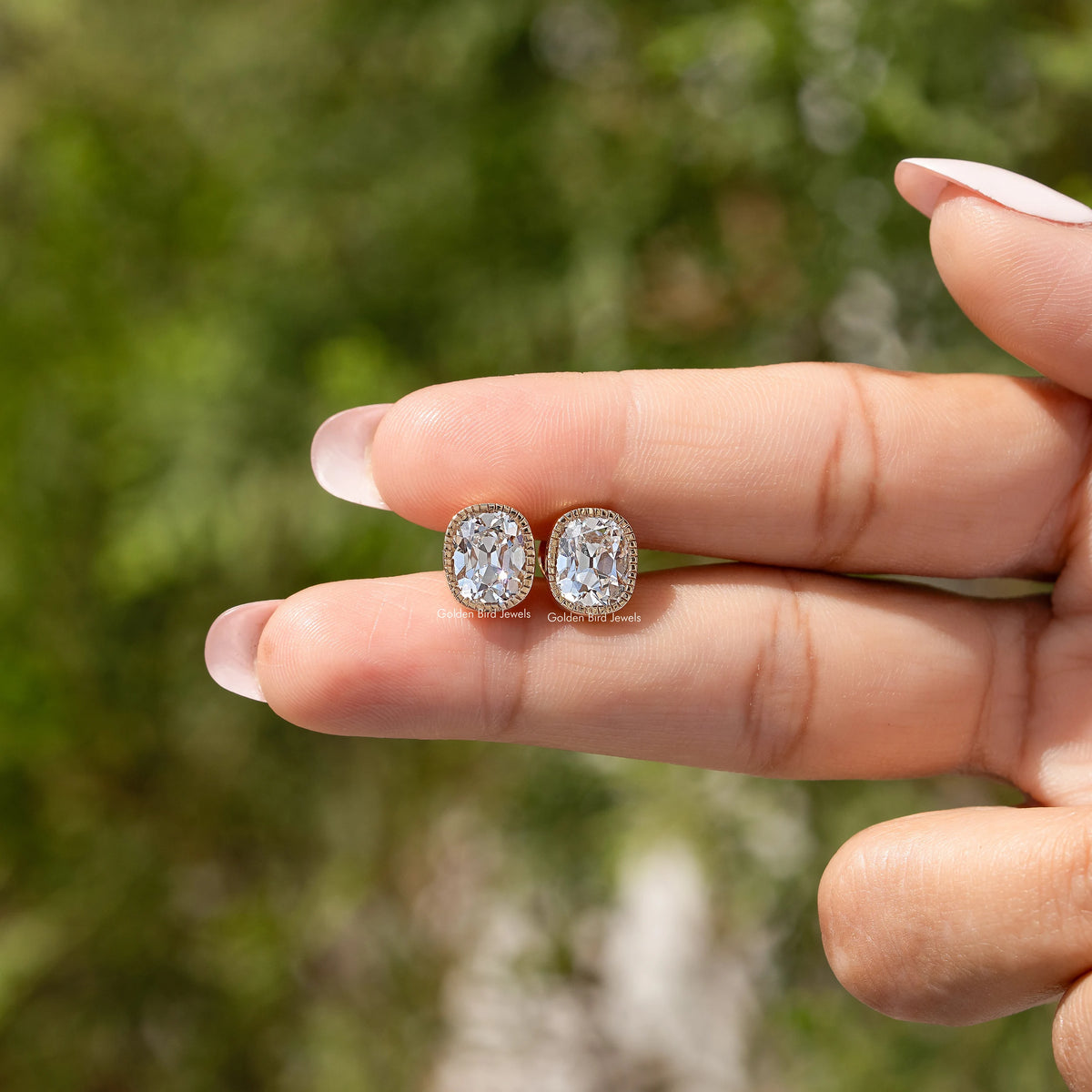 Cushion-cut diamond stud earrings with milgrain bezel in gold settings in hand