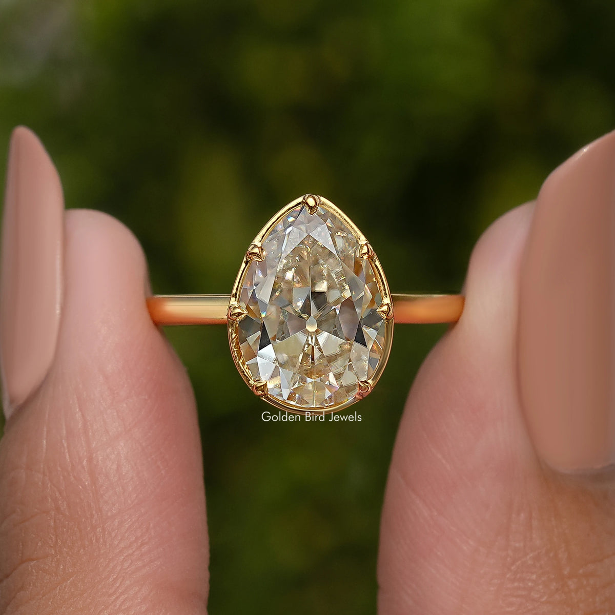 Gold-tone pear moissanite solitaire engagement ring held between fingertips