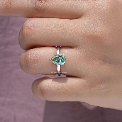 Pear-shaped moissanite engagement ring on fingertip, green center stone in prongs