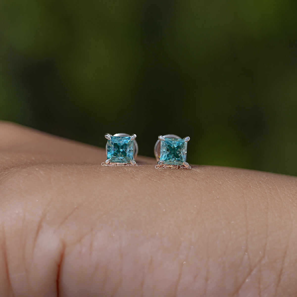 Two blue princess-cut screw-back stud earrings on skin close-up