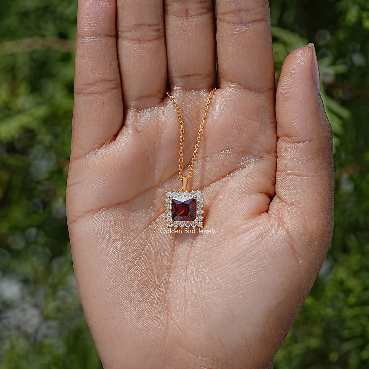 Square ruby-red gemstone pendant with halo stones, shown in open palm on gold chain